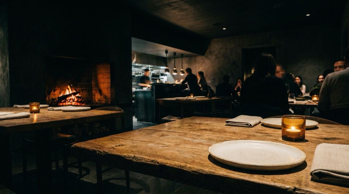 The BAD Studio build for BANKED: a dim dining room seen from a table, a wood fire burning high in a hearth at the left.